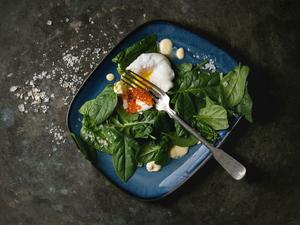 Poached egg on fresh spinach leaves with hollandaise sauce, red caviar served in blue square plate over old dark metal background. Top view, space. Vegetarian healthy eating