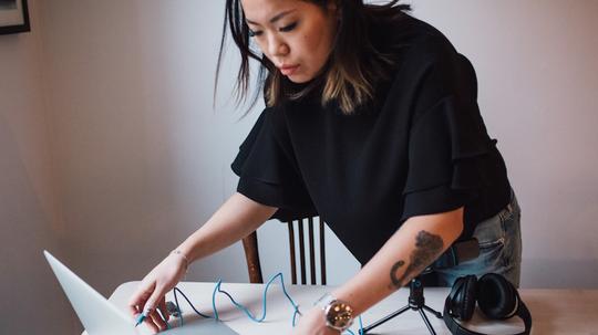 Female blogger using laptop standing by table with microphone at home