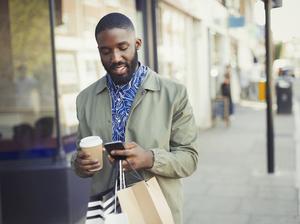 Young man with coffee and shopping bags texting with cell phone on urban sidewalk