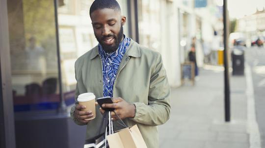 Young man with coffee and shopping bags texting with cell phone on urban sidewalk