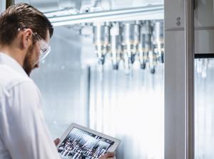 Man wearing lab coat and safety goggles at machine in factory looking at tablet