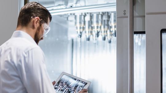 Man wearing lab coat and safety goggles at machine in factory looking at tablet