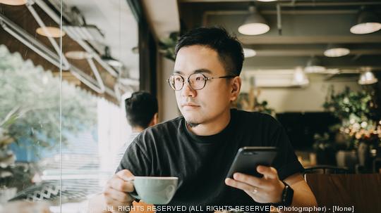 Smart young Asian man using smartphone and having coffee in cafe