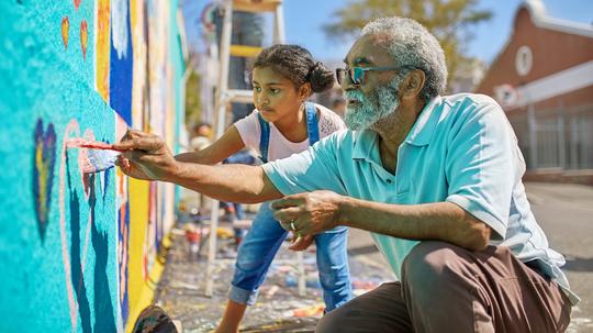 Grandfather and granddaughter volunteers painting vibrant mural on sunny urban wall