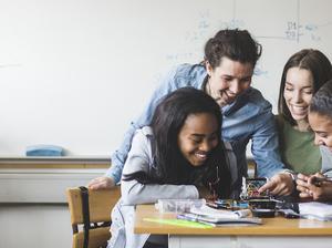 Smiling female teacher and high school teenage students preparing robot on desk in classroom