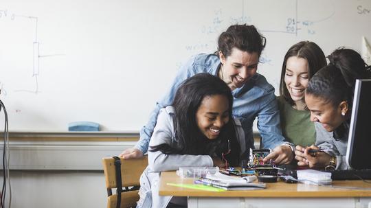 Smiling female teacher and high school teenage students preparing robot on desk in classroom