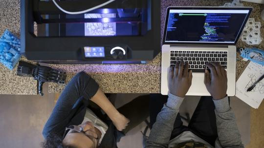 Overhead view designers working at laptop and using 3D printer in office