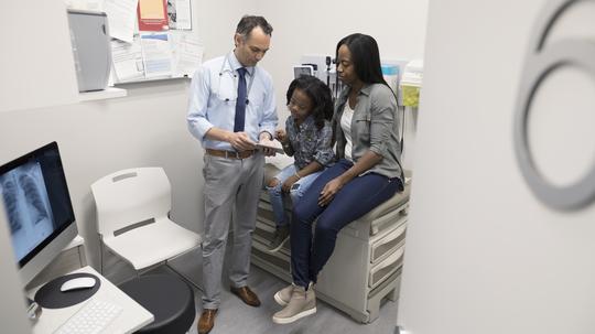 Male doctor with digital tablet talking to mother and daughter in clinic exam room