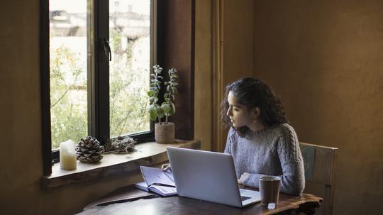 Young Woman of Mixed-ethnicity Works From Home Using Laptop Computer and Reference Book