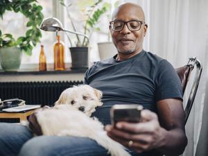 Smiling retired senior male using smart phone while sitting with dog in room at home