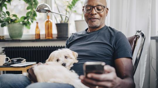 Smiling retired senior male using smart phone while sitting with dog in room at home