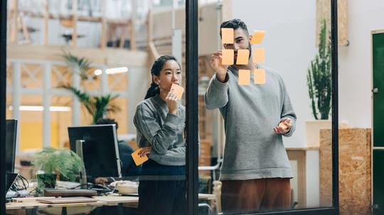 Colleagues discussing while sticking adhesive notes on glass wall in office