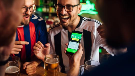 Friends drinking beer, watching soccer game and using mobile app for betting