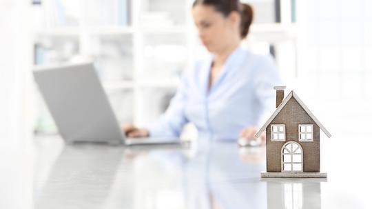 real estate office, home leaning on desk and woman agent working on computer in background