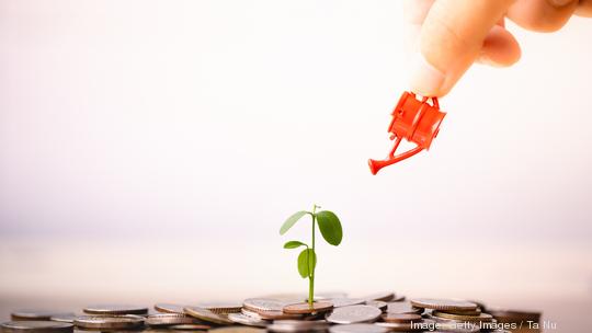 Woman hand holding red watering can with  money stack and seedling on top.