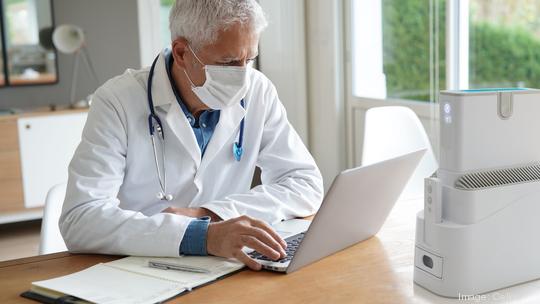 Doctor in office working on laptop, wearing protection mask