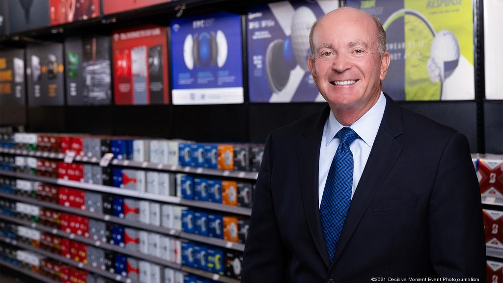 A Smiling Man In A Suit And Tie Standing Inside A Retail Store With Shelves Of Products And Promotional Displays Behind Him, Representing Leadership At Pga Tour Superstore. Who Owns Pga Superstore Detailed.