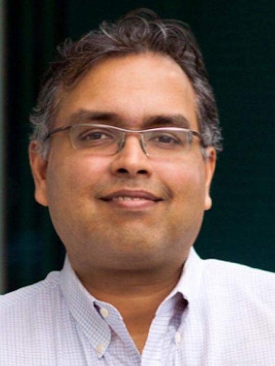 Auditoria CEO Rohit Gupta smiling wearing long-sleeved button-up shirt with his arms crossed leaning against a wall