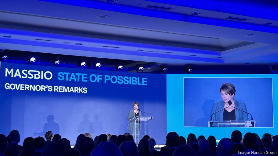 Governor Maura Healey on the stage at the MassBio State of Possible Conference. The screen behind her has the text Governor's Remarks and the entire room has a blue hue. 