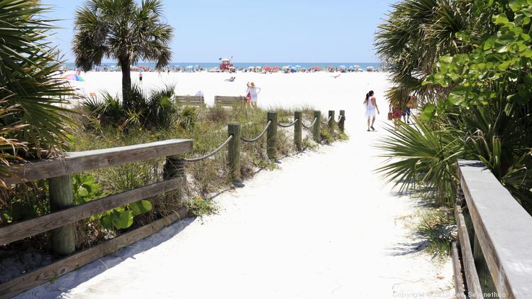 People entering Siesta Beach on Siesta Key through a sand covered wood walkway with wood railings surrounded by foliage on a sunny day.
